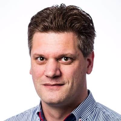 Een man met kort bruin haar en een gestreept overhemd kijkt naar de camera tegen een effen witte achtergrond. Hij belichaamt professionaliteit op het gebied van digitale forensische wetenschap en cyberbeveiliging.