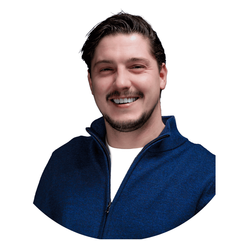 Een man met donker haar en een getrimde baard glimlacht, draagt een blauw ritsjack over een wit overhemd, tegen een effen witte achtergrond - perfect voor een algemeen profiel of een social engineering CTA.