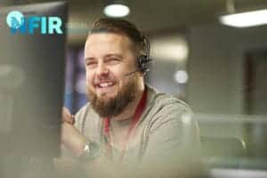 Man met baard draagt een headset en koord en glimlacht terwijl hij achter een bureau voor een computerscherm zit; het NFIR Social Engineering-logo, gericht op bewustwording van voice phishing, is zichtbaar in de linkerbovenhoek.